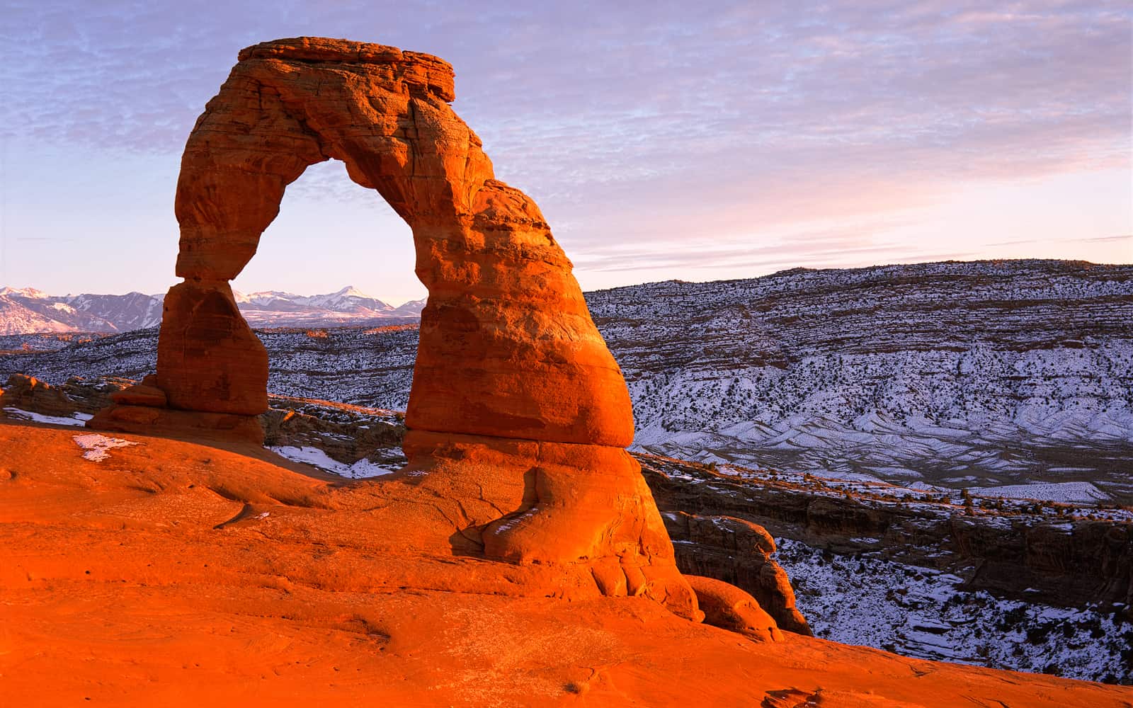Delicate Arch glowing red at sunset with snow-dusted cliffs behind it in Arches National Park, Utah